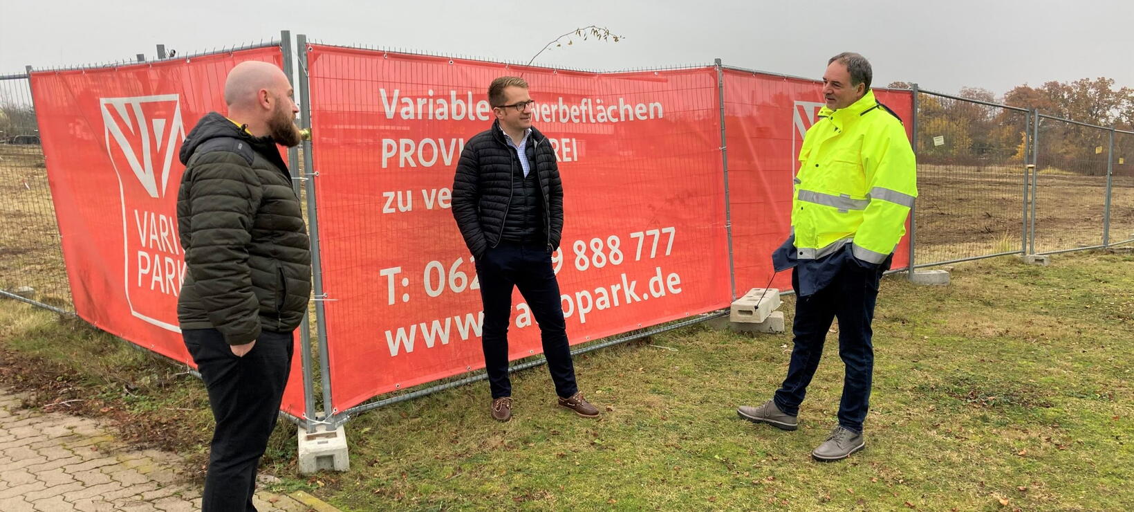 Drei Projektbeteiligte vor Banner-Werbung am Baufeld in Frankenthal